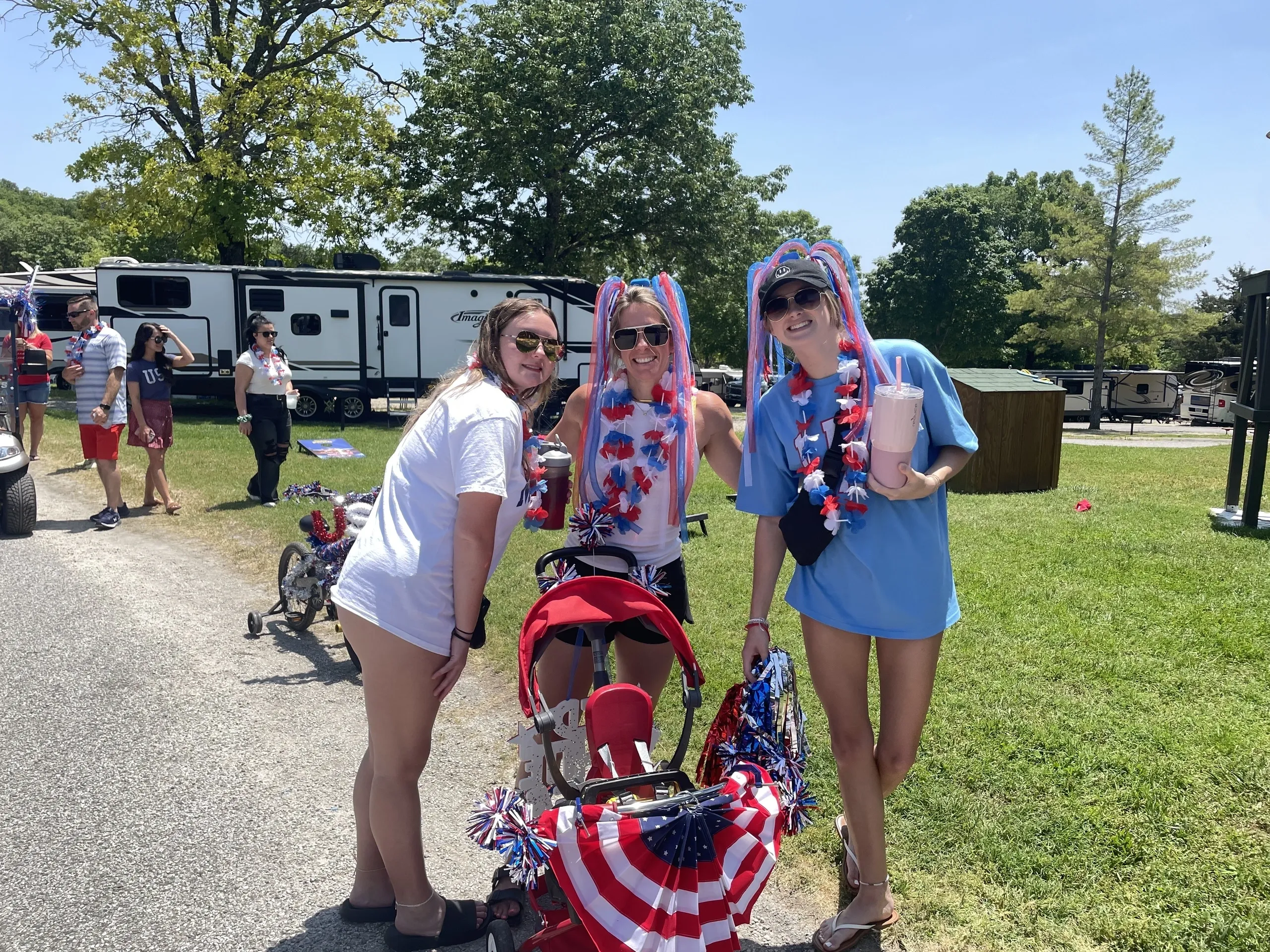 Three women are standing outside, wearing patriotic headbands and holding American flags