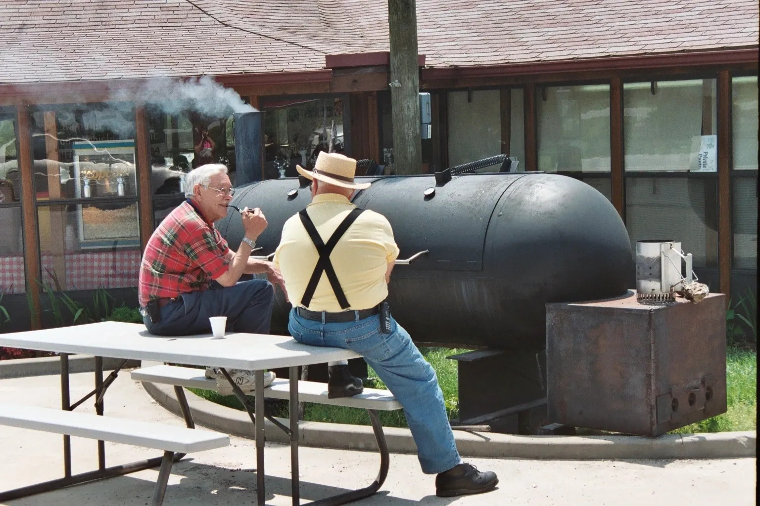 A man wearing a yellow shirt is sitting at a picnic table next to a large black grill